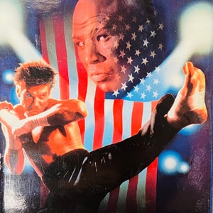 May include: A VHS tape cover for the movie Kickboxer 2. The cover features a man in a black martial arts uniform kicking another man in the face. The background is a red, white, and blue American flag. The text on the cover reads "Better Than Kickboxer" - LA Times and "Kickboxer 2".