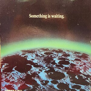 May include: VHS video cassette cover with a dark background and a close-up of a pair of green eyes. The text "The Dark Side of the Moon" is at the top of the cover. The text "Something is waiting." is in the middle of the cover. The bottom of the cover shows a view of the moon's surface. The Vidmark Entertainment logo is in the bottom left corner.