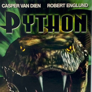 Pode incluir: Uma capa de VHS para o filme de terror "Python" com um close-up da cabeça de uma cobra com dentes afiados. A capa também apresenta os atores Casper Van Dien e Robert Englund. O texto "Sessenta pés de puro terror!" está impresso na capa.