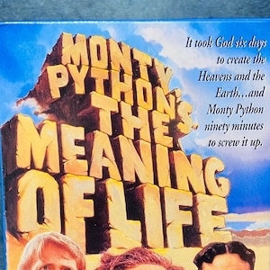 May include: A VHS video tape for the film "Monty Python's The Meaning of Life." The box has a colourful design with the film's title in large, blocky letters and images of the cast. The text on the box reads, "It took God six days to create the Heavens and the Earth...and Monty Python ninety minutes to screw it up."