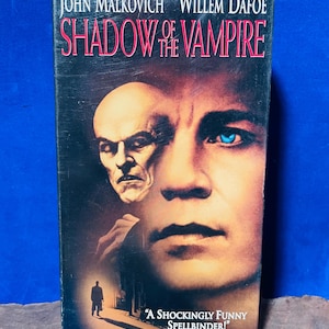 Puede incluir: Una cinta VHS de la película "Shadow of the Vampire" con John Malkovich y Willem Dafoe. La portada muestra dos caras, una calva y otra con ojos azules, con el título de la película en rojo. También es visible la cita "A SHOCKINGLY FUNNY SPELLBINDER!".