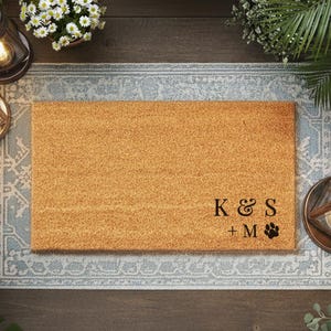 Op de afbeelding: Een rechthoekige kokosmat met de initialen "K & S" en "+ M" met een pootafdruk. De mat is omgeven door groen, bloemen en lantaarns, waardoor een uitnodigende buitenscène ontstaat.