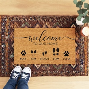 May include: A rectangular doormat with the words "welcome to our home" in black script. Below the text are paw prints, footprints, and names: Max, Kim, Noah, Tom, and Luna. The mat is on a patterned rug.