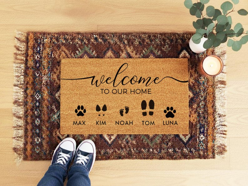 May include: A rectangular doormat with the words "welcome to our home" in black script. Below the text are paw prints, footprints, and names: Max, Kim, Noah, Tom, and Luna. The mat is on a patterned rug.