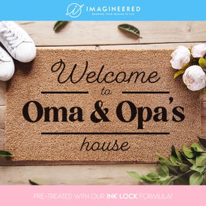 May include: A brown coir doormat with the text "Welcome to Oma & Opa's house" in black lettering. The doormat is on a wooden surface with white sneakers and white flowers.
