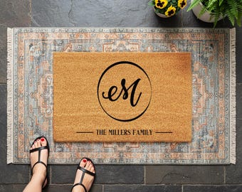 May include: A rectangular coir doormat with a black circular design containing the letters "eM" and the words "THE MILLERS FAMILY" in black. The mat is on a patterned rug. Black sandals are visible.
