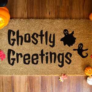 May include: A brown coir doormat with the text "Ghostly Greetings" in black lettering. Two black ghost silhouettes are on the doormat.