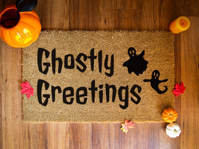 May include: A brown coir doormat with the text "Ghostly Greetings" in black lettering. Two black ghost silhouettes are on the doormat.