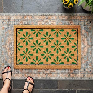 May include: A rectangular doormat with a floral pattern in green on a tan background. The doormat is placed on a patterned rug with a fringe. Black sandals are visible at the bottom of the image.