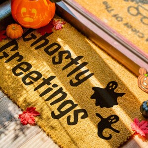 May include: A brown doormat with the text "Ghostly Greetings" in black lettering. The doormat is decorated with fall-themed items, including pumpkins, leaves, and ghosts.