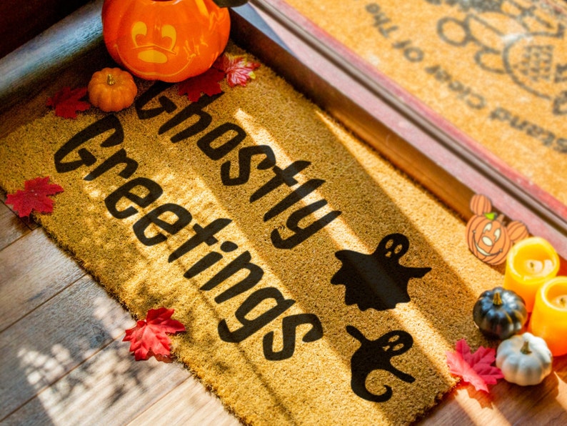 May include: A brown doormat with the text "Ghostly Greetings" in black lettering. The doormat is decorated with fall-themed items, including pumpkins, leaves, and ghosts.