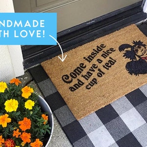 May include: A brown coir doormat with the text "Come inside and have a nice cup of tea!" and a cartoon illustration of a black and red creature. The doormat is on a black and white checkered surface.