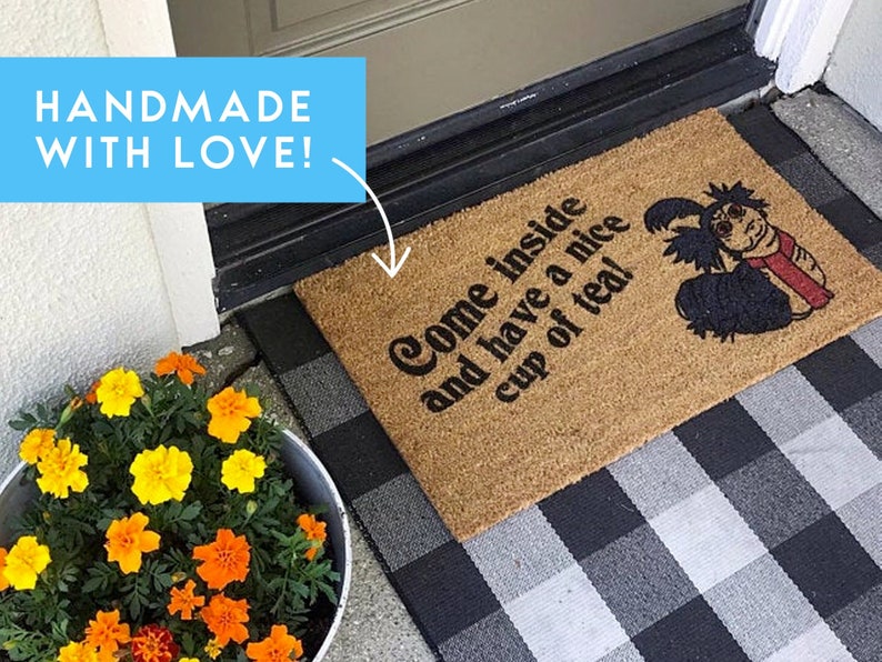May include: A brown coir doormat with the text "Come inside and have a nice cup of tea!" and a cartoon illustration of a black and red creature. The doormat is on a black and white checkered surface.