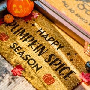 May include: A brown doormat with the text "Happy Pumpkin Spice Season" in black lettering. The doormat is decorated with fall-themed elements, including pumpkins, leaves, and stars.