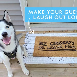 May include: A brown doormat with the text "Be groovy or leave, man" in black lettering. A husky dog with blue eyes is sitting on a wooden deck in front of the doormat.