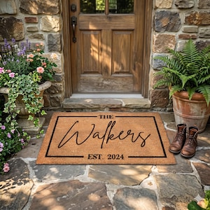 May include: A rectangular doormat with the text "THE Walkers EST. 2024" in black script. The mat is brown with a black border, placed in front of a wooden door. Brown leather boots are placed to the right of the mat.