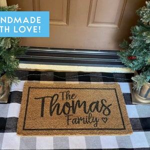 May include: A brown coir doormat with the text "The Thomas Family" in black cursive lettering. The doormat is on a black and white checkered rug in front of a brown door. There are two small potted Christmas trees on either side of the door.