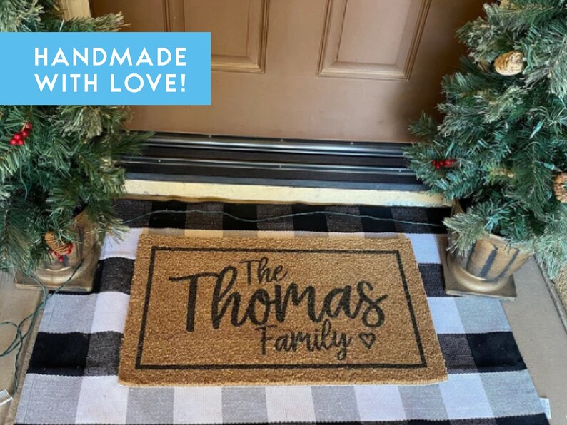 May include: A brown coir doormat with the text "The Thomas Family" in black cursive lettering. The doormat is on a black and white checkered rug in front of a brown door. There are two small potted Christmas trees on either side of the door.