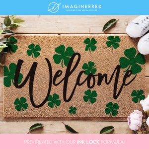 Puede incluir: Una alfombra rectangular de color marrón natural con la palabra "Welcome" en una fuente de escritura marrón oscuro, rodeada de tréboles verdes. El diseño está sobre un fondo de madera. Zapatillas blancas son visibles en la esquina superior derecha.