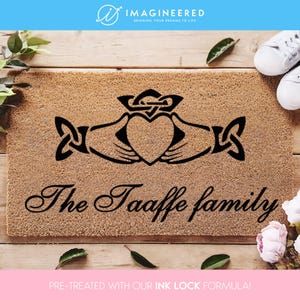 Könnte beinhalten: Eine braune Fußmatte mit einem schwarzen keltischen Knotendesign, das zwei Hände zeigt, die ein Herz umschließen. Der Text "The Taaffe family" ist in einer kursivschriftlichen Schrift unter dem Design geschrieben.