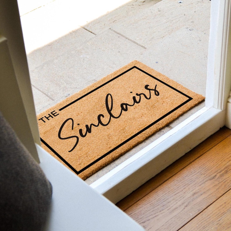 May include: A rectangular coir doormat with a black border and the family name "The Sinclair's" in a cursive font. The mat is placed in a doorway, with a view of the floor and door frame.