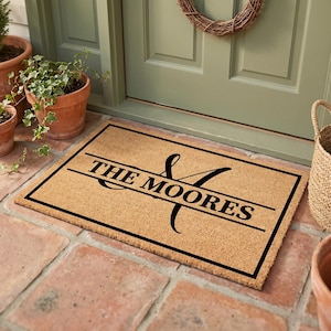 May include: A rectangular doormat made of natural coir fiber. The mat features a black border and the words "THE MOORES" in a bold, black font, with a decorative ampersand and initial. The mat is placed on a brick patio.