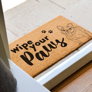 Pu&ograve; includere: Un tappetino per porta in fibra di cocco marrone con la scritta "Wipe your paws" e un'illustrazione in bianco e nero di un bulldog francese.
