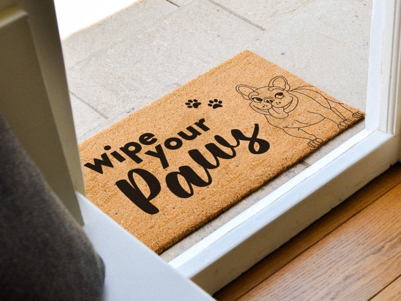 Pu&ograve; includere: Un tappetino per porta in fibra di cocco marrone con la scritta "Wipe your paws" e un'illustrazione in bianco e nero di un bulldog francese.