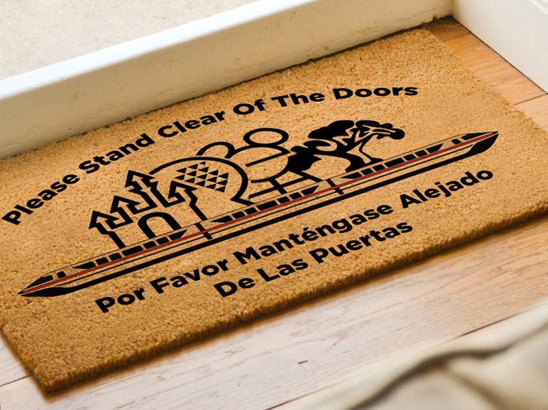 May include: A brown coir doormat with the text "Please Stand Clear Of The Doors" and "Por Favor Mant&eacute;ngase Alejado De Las Puertas" in black lettering. The doormat features a black silhouette of a monorail train, a castle, and a tree.