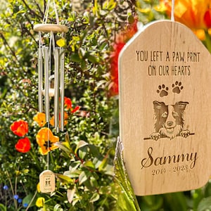 May include: A wooden wind chime with a memorial plaque. The plaque has the text "YOU LEFT A PAW PRINT ON OUR HEARTS" and a dog illustration. The wind chime is hanging in a garden with orange and yellow flowers.