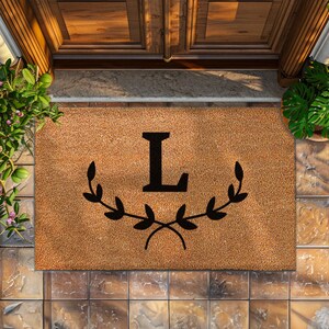 May include: A brown coir doormat with the letter "L" in black surrounded by two black leafy branches.