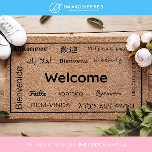 Puede incluir: Una alfombra de coco marrón con la palabra "Welcome" en el centro, rodeada de la palabra "Welcome" en diferentes idiomas, incluyendo español, francés, alemán, italiano, polaco, árabe, hindi y japonés. La alfombra está pretratada con una fórmula de bloqueo de tinta.