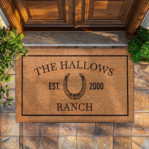 Puede incluir: Una alfombra de coco marrón con texto negro que dice "The Hallows Ranch Est. 2000" y un diseño de herradura negro.