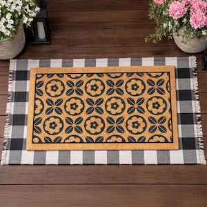 May include: A rectangular doormat with a floral pattern in black on a tan background. The doormat is placed on a black and white checkered rug with fringed edges. The scene is set on a wooden deck, with potted plants and lanterns in the background.