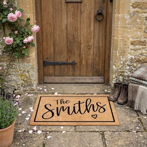 May include: A rectangular doormat with a black border and the name "The Smiths" in a cursive font, with a heart symbol. The mat is made of a natural fiber material and is placed in front of a wooden door.