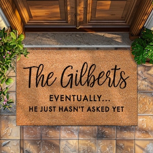 Puede incluir: Una alfombra de coco marrón con el texto "The Gilberts" en fuente cursiva negra. Debajo del nombre, se lee "Eventually... He just hasn't asked yet."