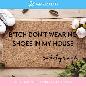 Puede incluir: Felpudo rectangular marrón con el texto "B*TCH DON'T WEAR NO SHOES IN MY HOUSE - roddy ricch" en negro. Zapatillas blancas y decoraciones florales en el fondo.