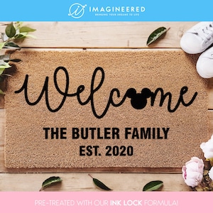 May include: A brown coir doormat with the text "Welcome" in black cursive lettering. A black Mickey Mouse head is incorporated into the word "Welcome". The text "The Butler Family Est. 2020" is also in black lettering.
