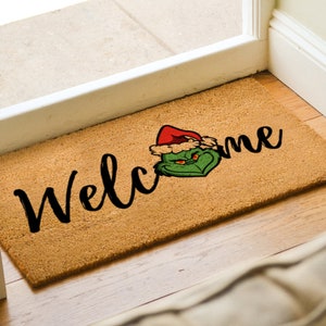 May include: A brown coir doormat with the word "Welcome" in black cursive lettering. A green Grinch character wearing a red Santa hat is positioned to the right of the word "Welcome".