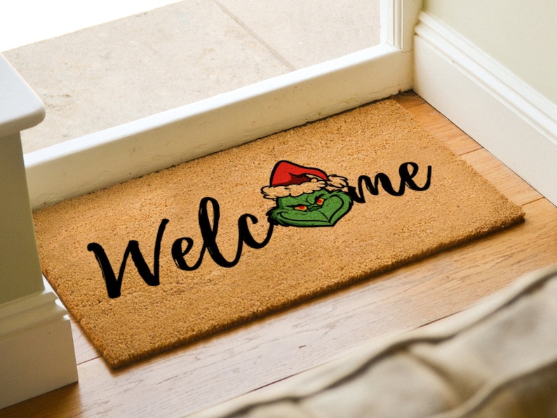 May include: A brown coir doormat with the word "Welcome" in black cursive lettering. A green Grinch character wearing a red Santa hat is positioned to the right of the word "Welcome".
