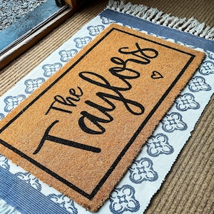 May include: A rectangular coir doormat with a black border and the words "The Taylors" in a cursive font. A small heart is next to the last name. The doormat is on top of a patterned rug with a blue and white design.