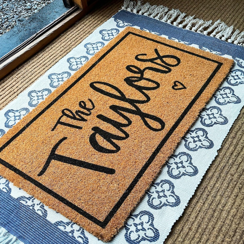 May include: A rectangular coir doormat with a black border and the words "The Taylors" in a cursive font. A small heart is next to the last name. The doormat is on top of a patterned rug with a blue and white design.