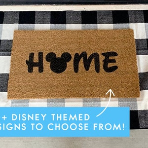 May include: A brown coir doormat with the word "HOME" in black lettering. The "O" is replaced with a black Mickey Mouse head. The doormat is on top of a black and white checkered rug. The text "100+ DISNEY THEMED DESIGNS TO CHOOSE FROM!" is below the doormat.