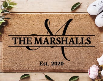 May include: A brown coir doormat with the text "THE MARSHALLS" and "Est. 2020" in black lettering. The text is in a decorative font with a stylized "M" monogram.