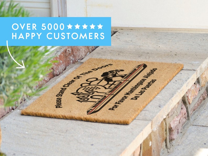 May include: A brown coir doormat with the text "Please Stand Clear of the Doors" and "Por Favor Mant&eacute;ngase Alejado De Las Puertas" in black text. The doormat features a stylised image of a monorail.