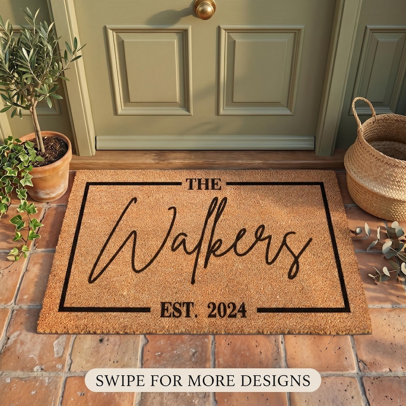 May include: A rectangular coir doormat with a black border and the text "THE Walkers EST. 2024" in a cursive font. The mat is placed in front of a green door, with a potted plant and a woven basket nearby.