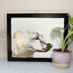 May include: A framed art print featuring a gold foil dog portrait on a white background. The black frame measures 10 inches wide and 8 inches tall. A potted plant with green leaves sits to the right of the artwork.