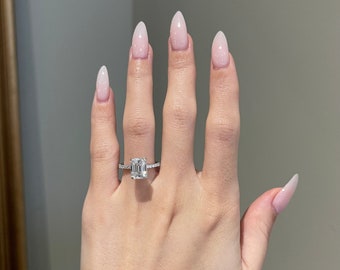 Puede incluir: Un anillo de compromiso plateado con un gran diamante de talla esmeralda. El anillo está engastado con diamantes más pequeños a lo largo de la banda. La mano tiene uñas largas de color rosa claro.