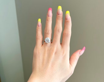 May include: A close-up of a hand wearing a gold ring with a large, round, clear diamond. The fingernails are painted with a yellow and pink ombre design.