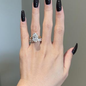 May include: A hand displaying two gold rings. The primary ring features a large, oval-cut diamond. The second ring is a gold band with smaller diamonds. The nails are painted black with a starry design.